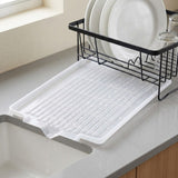 A white plastic under-sink drying rack with slotted drain holes sits on a gray kitchen countertop next to a sink, with a black wire dish rack holding white plates in the background.