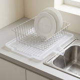 A clear plastic dish drainer tray with a silver metal wire rack holding white plates, placed on a kitchen counter next to a sink with running water.