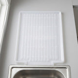 A white plastic sink drying rack with a slotted grid design, shown hanging above a stainless steel sink.