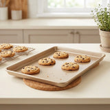 A rectangular beige baking sheet resting on a round cork trivet on a white kitchen countertop, holding several chocolate chip cookies. A cooling rack with more cookies is visible in the background, along with a potted plant and soft kitchen lighting.