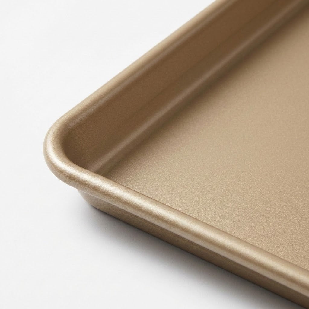 Close-up of a rectangular baking pan or tray corner with rounded edges, shown in a warm metallic gold finish on a clean white background.