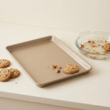A rectangular metal baking sheet with rounded corners in a warm beige finish, shown on a light cream countertop with cookies and crumbs, alongside a glass pie dish with residue.