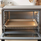 A close-up of a countertop convection oven with the door open, showing a metal baking tray on the interior rack in a kitchen setting.
