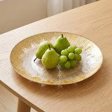 A round glass plate with a textured gold rim sits on a light wood table, holding three green pears and a small bunch of green grapes.