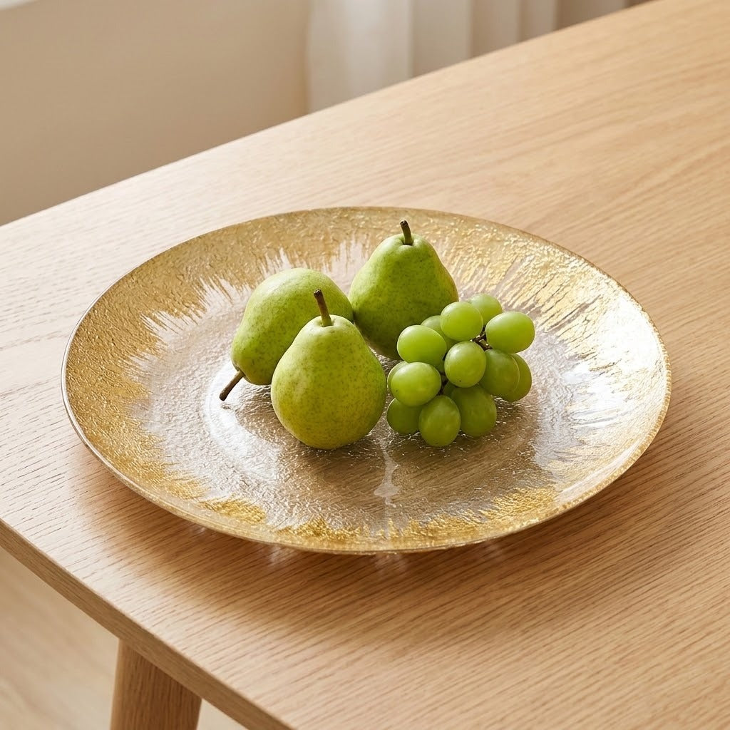 A round glass plate with a textured gold rim sits on a light wood table, holding three green pears and a small bunch of green grapes.
