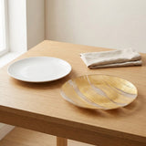 A round white ceramic plate and a round gold plate with a brushed metallic finish sit on a light wood table next to a folded beige cloth napkin in a bright room.