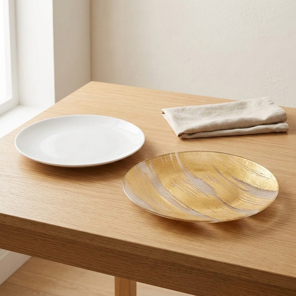 A round white ceramic plate and a round gold plate with a brushed metallic finish sit on a light wood table next to a folded beige cloth napkin in a bright room.