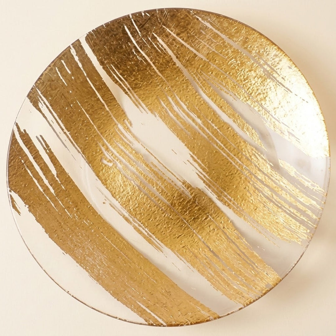 A round decorative plate featuring an abstract gold brushstroke pattern on a white background, photographed from directly above against a light cream backdrop.