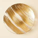 A round decorative plate featuring a textured gold and white abstract brushstroke pattern, photographed from directly above on a light beige background.