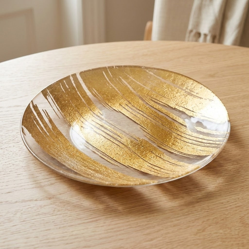 A round glass plate with an abstract gold leaf pattern, resting on a light wood table.