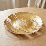 A round glass plate with an abstract gold leaf pattern, resting on a light wood table.