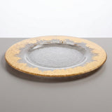 A round glass charger plate with a textured clear center and a wide gold-rimmed border, shown on a neutral gray surface against a white background.