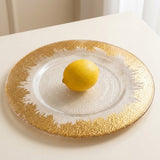A round glass charger plate with a textured gold rim and white center, holding a yellow lemon on a light beige tabletop.