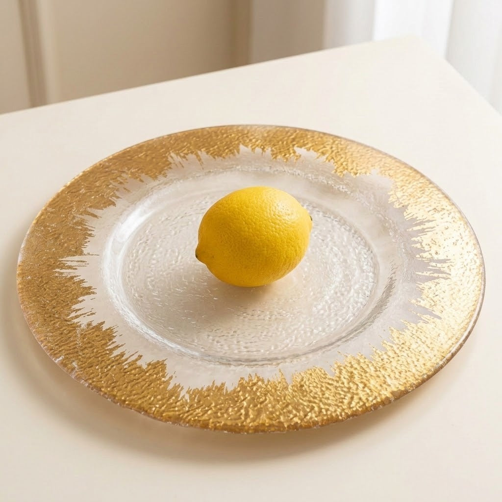 A round glass charger plate with a textured gold rim and white center, holding a yellow lemon on a light beige tabletop.
