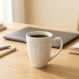 A white ceramic coffee mug with vertical fluted detailing sits on a light wood desk, filled with black coffee, with a blurred laptop and stationery in the background.