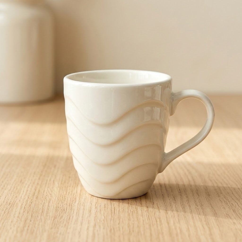A white ceramic coffee mug with a wavy, ribbed pattern and a curved handle, sitting on a light wood tabletop with a soft-focus beige background.