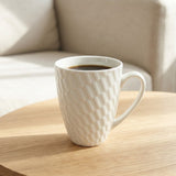 A white ceramic mug with a textured diamond pattern, filled with black coffee and sitting on a light wood table in a softly lit room.