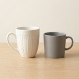 Two ceramic mugs on a light wood tabletop against a warm beige background. The mug on the left is off-white with a ribbed vertical texture and a curved handle.