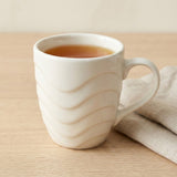 A white ceramic mug with a wavy, ribbed exterior pattern, filled with tea and resting on a light wood table next to a beige linen napkin.