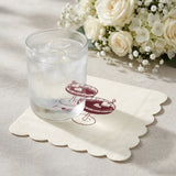 A clear glass tumbler filled with ice sits on a scalloped cream napkin printed with a burgundy cocktail glass illustration. The napkin is placed on a light tabletop with white flowers blurred in the background.