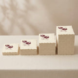 Four stacks of cream-colored paper napkins with scalloped edges, each printed with a small illustration of two coffee cups and the word 'Cappuccino', arranged in ascending sizes on a light beige tablecloth.