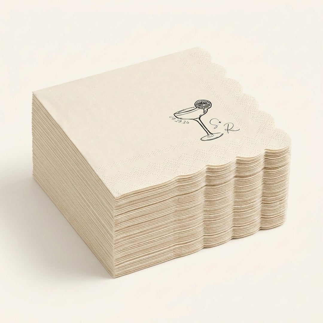 A stack of square paper napkins in a warm cream color, featuring a scalloped edge and a black printed illustration of a cocktail glass with a cherry on top, set against a solid cream background.