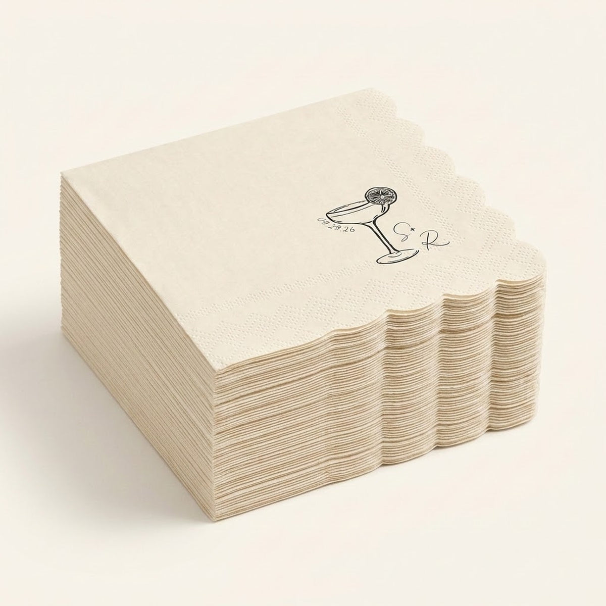 A stack of square paper napkins in a warm cream color, featuring a scalloped edge and a black printed illustration of a cocktail glass with a cherry on top, set against a solid cream background.