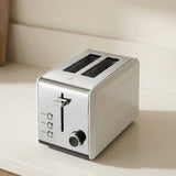 A modern two-slot toaster with a brushed silver metallic finish, featuring a sliding lever, two slots, and a round dial, shown on a light beige countertop.