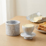 A pair of light gray marble butter dishes on a light wood table, with one dish open and the other holding a pat of butter. A plate with toast and an unwrapped stick of butter sits in the softly blurred background.