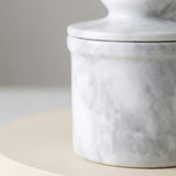 A close-up of a white and gray marble lidded container or jar sitting on a light beige surface against a soft gray background.