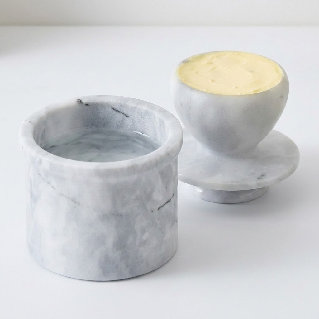 A pair of light gray marble butter dishes on a white background, one showing the pale yellow butter inside and the other empty.