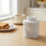 A white marble butter dish with a matching lid sits on a light wood table in a bright kitchen setting, with a cup of coffee, toast, and a knife blurred in the background.