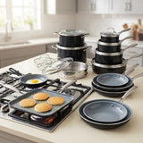 A multi-piece nonstick cookware set arranged on a kitchen countertop, including black and gray pots with lids, stainless steel pots, frying pans, a skillet with pancakes cooking on a gas stove, and glass lids. A real kitchen environment is visible in the background.