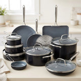 A 16-piece cookware set in matte dark gray with stainless steel handles and trim, displayed on a light beige countertop in a bright, modern kitchen setting.