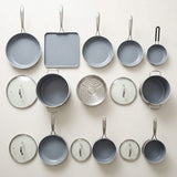 A set of light gray nonstick cookware pieces arranged on a light beige background, including multiple frying pans, a square baking pan, saucepans, lids, a steamer insert, and a small hanging utensil.