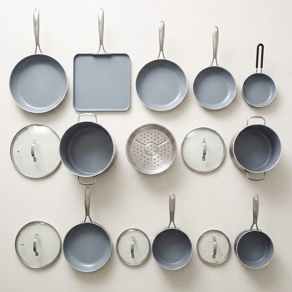 A set of light gray nonstick cookware pieces arranged on a light beige background, including multiple frying pans, a square baking pan, saucepans, lids, a steamer insert, and a small hanging utensil.