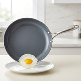 A dark gray non-stick frying pan with a stainless steel handle, shown tilted over a white plate with a fried egg resting on the rim, set against a softly blurred kitchen background.