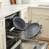 Gray nonstick cookware, including a saucepan and frying pan, loaded into a stainless steel dishwasher inside a bright kitchen.