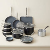 A cookware set arranged on a light beige countertop against a matching neutral wall. The set includes dark gray nonstick pots, pans, lids, a rectangular baking pan, a steamer insert, and a small silver saucepan with a black handle.