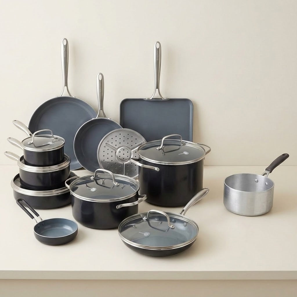 A cookware set arranged on a light beige countertop against a matching neutral wall. The set includes dark gray nonstick pots, pans, lids, a rectangular baking pan, a steamer insert, and a small silver saucepan with a black handle.