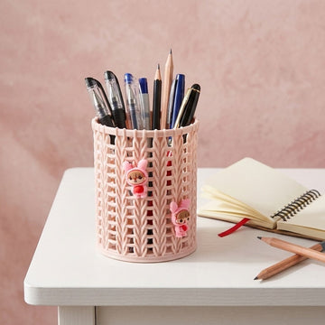 A pink plastic pencil holder with a knitted pattern and two small cartoon charms, filled with pens and pencils, sitting on a white desk next to an open notebook.