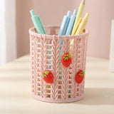 A pink plastic pencil holder with a knitted pattern and three 3D strawberry decorations, filled with pastel-colored pens and pencils, set on a light beige surface against a soft pink background.