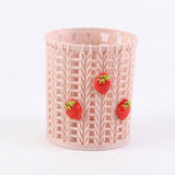 A light pink cylindrical basketweave-patterned cup or candle holder decorated with three small 3D strawberry embellishments, shown on a plain white background.
