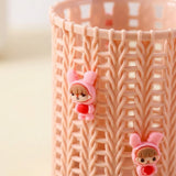 A close-up of a peach-pink plastic cylindrical holder with a knitted cutout pattern, decorated with two small cartoon character charms wearing pink bunny hats and red hearts.