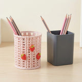 Two pencil holders on a light wood desk against a cream wall. The left holder is a pink knitted-texture cup with three red strawberry decorations, holding three pencils.