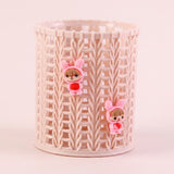 A cylindrical plastic basket with a cream-colored interlocking wavy pattern, decorated with two small pink bunny-ear character charms holding red hearts.