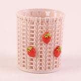 A cylindrical plastic cup or tumbler with a knitted pattern in a very pale pink, decorated with three small 3D strawberry embellishments on the front, set against a solid pale pink background.