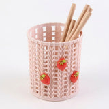 A light pink plastic pencil holder with a knitted texture, decorated with three small red strawberries, containing wooden pencils on a white background.