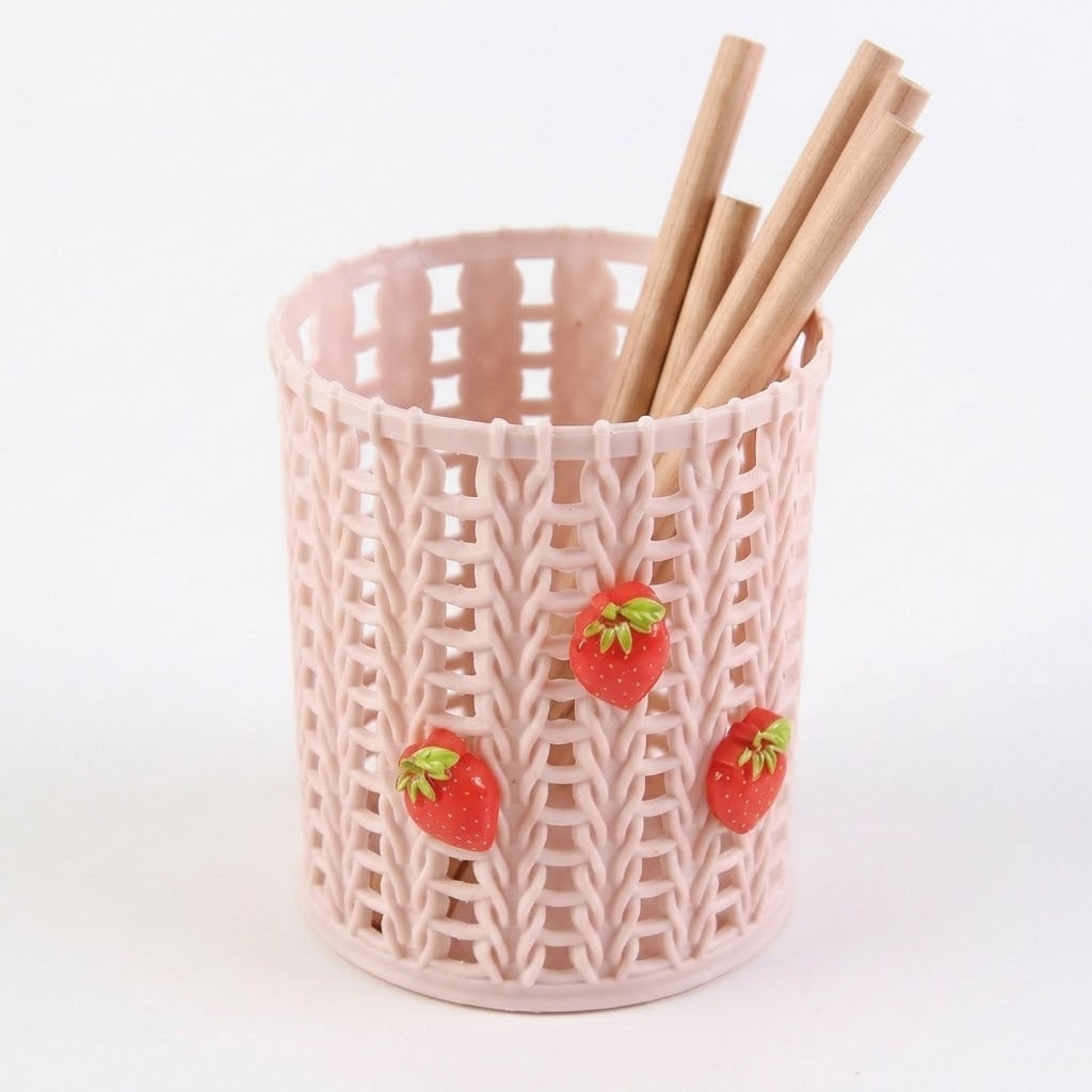 A light pink plastic pencil holder with a knitted texture, decorated with three small red strawberries, containing wooden pencils on a white background.