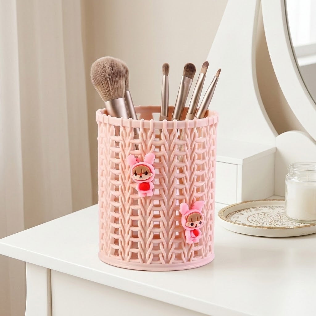 A pink plastic makeup brush holder with a knitted pattern and two small cartoon character decorations, holding several makeup brushes on a white vanity table.